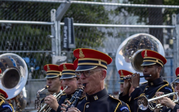 A U.S. Army marching band performs in what appears to be a parade or public event. The musicians are dressed in formal ceremonial uniforms featuring dark navy jackets with gold trim, blue trousers, and distinctive red-topped caps. The foreground features a clarinetist with visible rank insignia and service medals, surrounded by bandmates playing brass instruments including tubas and trumpets.