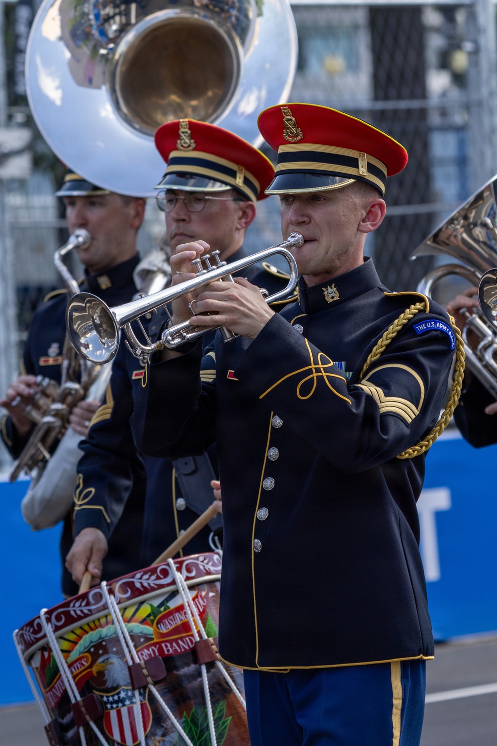 Members of The U.S. Army Band perform in a parade or public event, wearing formal dress uniforms with distinctive navy blue and gold trim and red-topped caps. The foreground musician plays a silver trumpet while others behind him play additional brass instruments and a decorated bass drum. The drum bears markings identifying it as belonging to 'The U.S. Army Band' based in Washington.