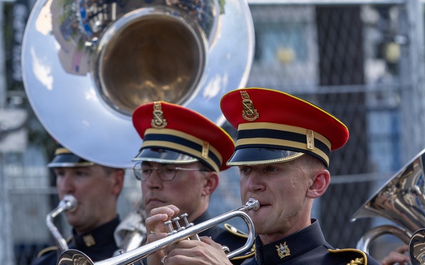 Members of The U.S. Army Band perform in a parade or public event, wearing formal dress uniforms with distinctive navy blue and gold trim and red-topped caps. The foreground musician plays a silver trumpet while others behind him play additional brass instruments and a decorated bass drum. The drum bears markings identifying it as belonging to 'The U.S. Army Band' based in Washington.