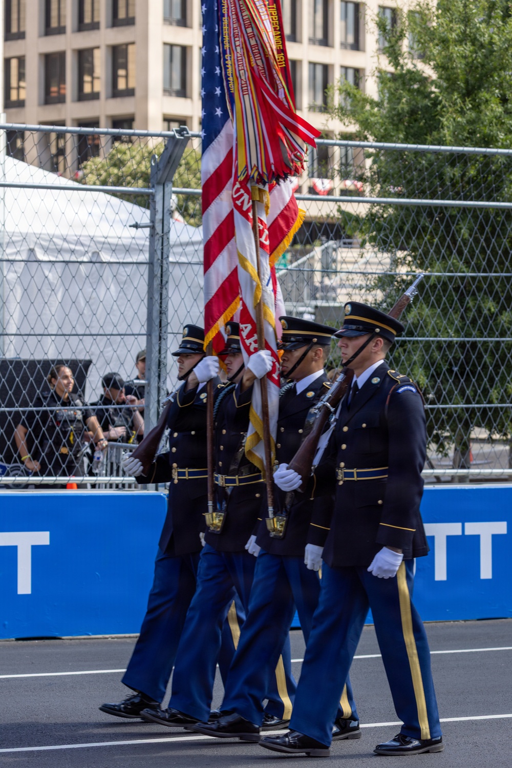A U.S. Army Color Guard unit is marching in formal dress uniform during what appears to be a ceremonial parade or public event. Four soldiers are carrying the U.S. national flag and what appears to be a unit guidon/colors with battle streamers, while two armed guards flank them with rifles. The event takes place on a closed street with crowd barriers and spectators visible in the background.