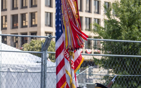 A U.S. Army Color Guard unit is marching in formal dress uniform during what appears to be a ceremonial parade or public event. Four soldiers are carrying the U.S. national flag and what appears to be a unit guidon/colors with battle streamers, while two armed guards flank them with rifles. The event takes place on a closed street with crowd barriers and spectators visible in the background.
