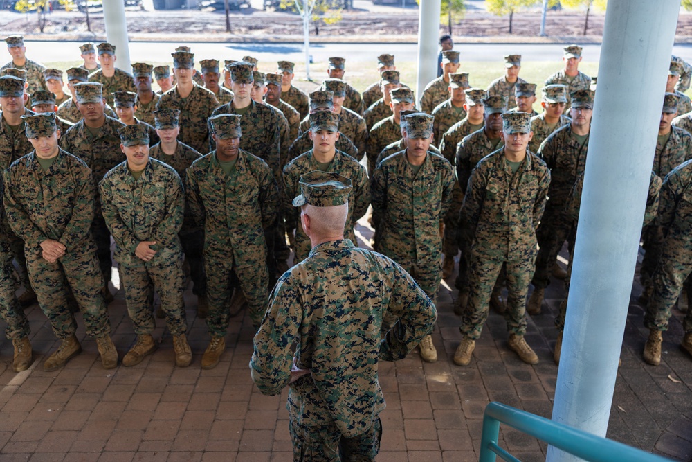 A senior military officer addresses a large formation of uniformed service members standing at attention outdoors. The personnel are wearing MARPAT (Marine Pattern) digital camouflage uniforms and covers, consistent with United States Marine Corps dress. The setting appears to be a paved outdoor area on a military installation.
