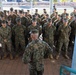 A senior military officer addresses a large formation of uniformed service members standing at attention outdoors. The personnel are wearing MARPAT (Marine Pattern) digital camouflage uniforms and covers, consistent with United States Marine Corps dress. The setting appears to be a paved outdoor area on a military installation.