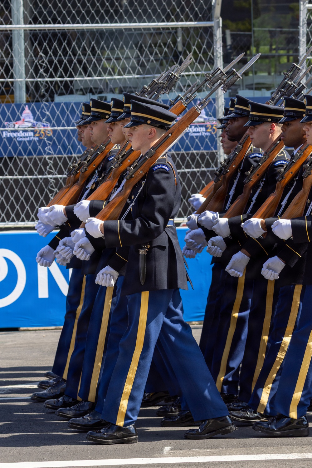 A U.S. Army Honor Guard unit is performing a ceremonial drill march, with soldiers in formal dress uniforms carrying bayonet-fixed rifles at shoulder arms. The event appears to be associated with the 'Freedom 250 Grand Prix' based on visible signage in the background. The soldiers are marching in precise formation on a paved surface, consistent with a public ceremonial or pre-race event.