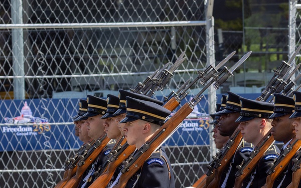 A U.S. Army Honor Guard unit is performing a ceremonial drill march, with soldiers in formal dress uniforms carrying bayonet-fixed rifles at shoulder arms. The event appears to be associated with the 'Freedom 250 Grand Prix' based on visible signage in the background. The soldiers are marching in precise formation on a paved surface, consistent with a public ceremonial or pre-race event.