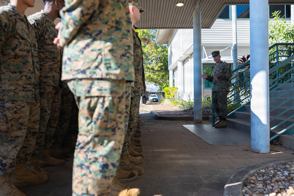 A group of military personnel in MARPAT (Marine Pattern) camouflage uniforms are standing in formation under a covered walkway of a building. A single service member, likely a senior officer or NCO, stands apart near a staircase and appears to be addressing the group. The setting appears to be a military installation, possibly in a tropical or subtropical location based on the vegetation visible.