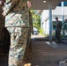 A group of military personnel in MARPAT (Marine Pattern) camouflage uniforms are standing in formation under a covered walkway of a building. A single service member, likely a senior officer or NCO, stands apart near a staircase and appears to be addressing the group. The setting appears to be a military installation, possibly in a tropical or subtropical location based on the vegetation visible.