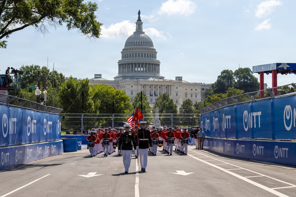 A U.S. Marine Corps marching band performs on what appears to be a temporary street circuit race track in front of the United States Capitol building in Washington, D.C. The track is lined with blue barrier walls bearing 'NTT' sponsor branding, suggesting this is an IndyCar or Formula E racing event. Marines in dress uniforms — both black and red — march in formation carrying the American flag and a Marine Corps flag.