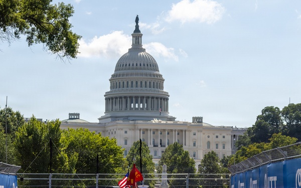 A U.S. Marine Corps marching band performs on what appears to be a temporary street circuit race track in front of the United States Capitol building in Washington, D.C. The track is lined with blue barrier walls bearing 'NTT' sponsor branding, suggesting this is an IndyCar or Formula E racing event. Marines in dress uniforms — both black and red — march in formation carrying the American flag and a Marine Corps flag.