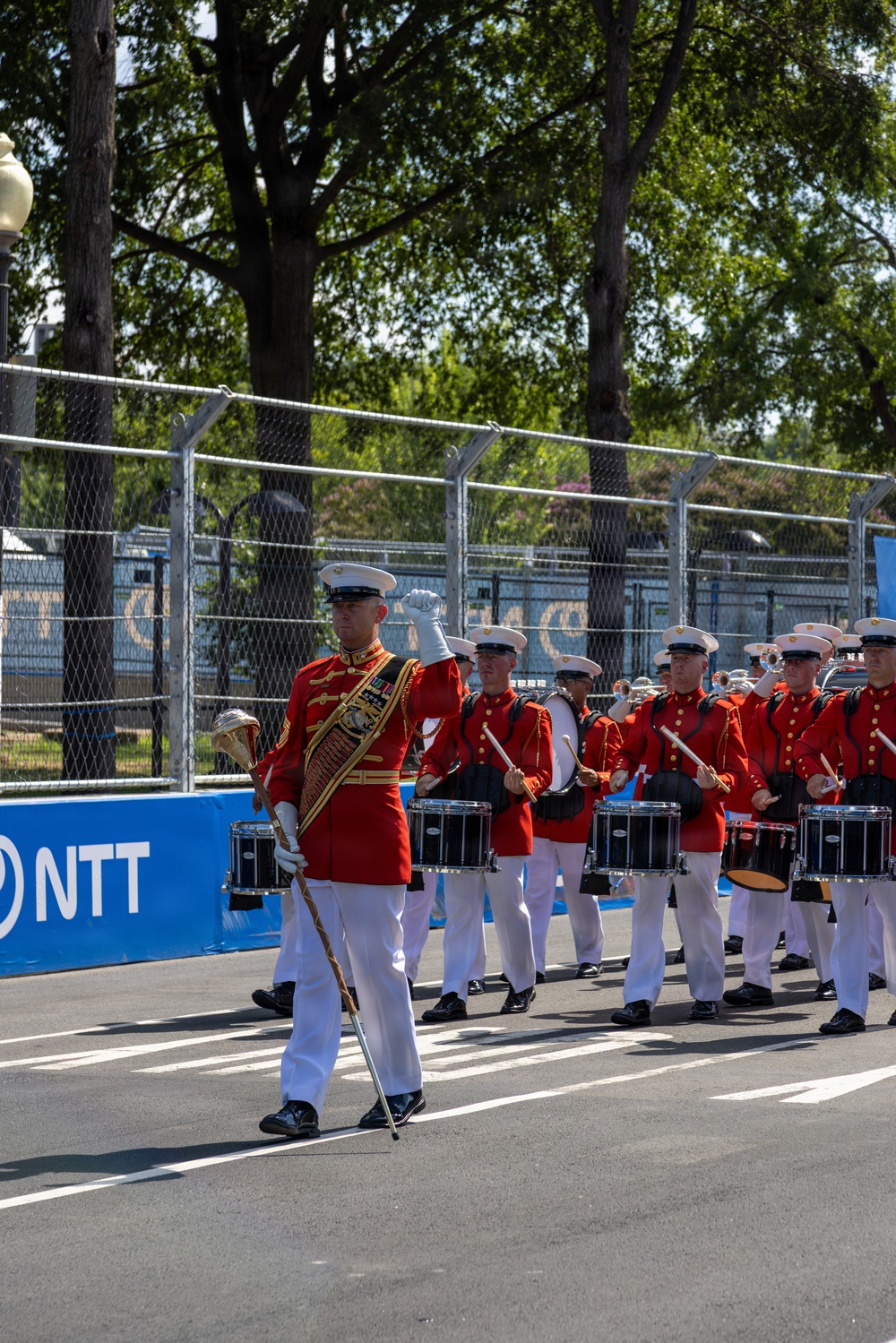 A United States Marine Corps marching band performs on what appears to be a racing circuit, identifiable by the 'NTT' sponsor banner and track markings. The band members are dressed in their distinctive scarlet and gold dress uniforms with white covers (caps) and white trousers. A drum major leads the procession, carrying a ceremonial mace, while drummers march in formation behind him.