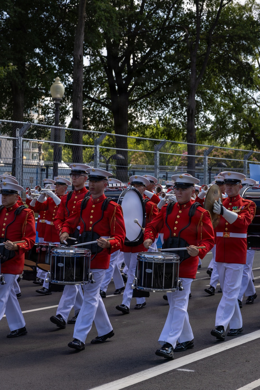 A military marching band, identifiable as the United States Marine Corps Band by their distinctive red tunics with gold buttons, white trousers, and white peaked caps, is performing in a parade. The musicians are playing snare drums, a bass drum, cymbals, and brass instruments while marching in formation on a paved road. The setting appears to be an outdoor urban environment with chain-link fencing and large trees visible in the background.