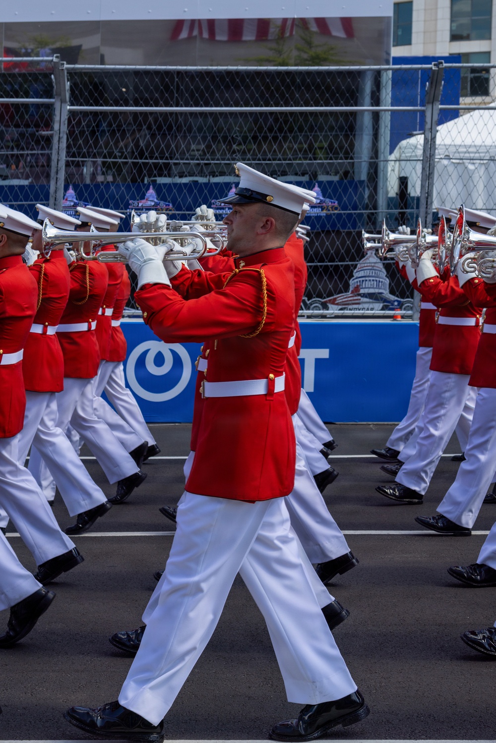 A military marching band, dressed in distinctive red tunics and white trousers with white caps, performs at what appears to be an IndyCar racing event. The musicians are playing brass instruments, including trumpets and French horns, while marching in formation. Signage in the background references 'IndyCar' and shows imagery of the U.S. Capitol, suggesting this may be the IndyCar Grand Prix on the Streets of Washington D.C. or a similar patriotic motorsport event.
