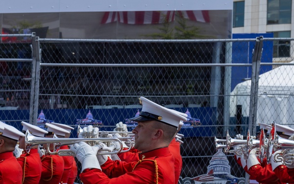 A military marching band, dressed in distinctive red tunics and white trousers with white caps, performs at what appears to be an IndyCar racing event. The musicians are playing brass instruments, including trumpets and French horns, while marching in formation. Signage in the background references 'IndyCar' and shows imagery of the U.S. Capitol, suggesting this may be the IndyCar Grand Prix on the Streets of Washington D.C. or a similar patriotic motorsport event.