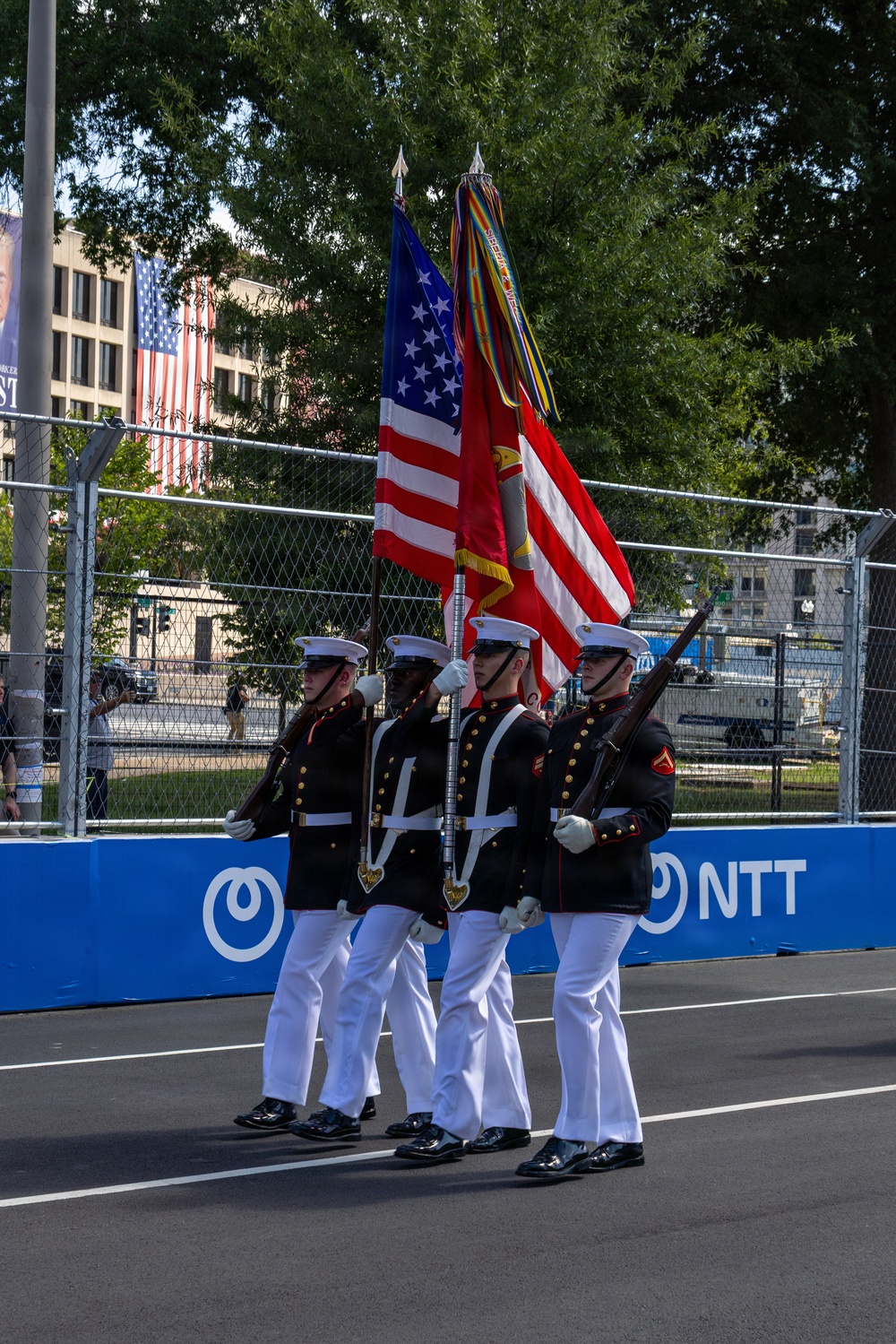 A U.S. Marine Corps Color Guard is marching in formation on a paved surface, carrying the American flag and what appears to be the Marine Corps flag along with a streamer-adorned staff. The Marines are dressed in their distinctive dress blues uniform with white trousers, white gloves, and white covers (caps). The event appears to take place at a motorsport venue, indicated by the NTT-branded barrier walls and chain-link fencing typical of street circuit racing.