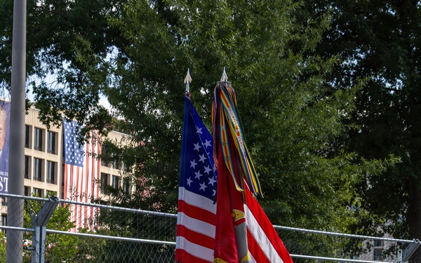 A U.S. Marine Corps Color Guard is marching in formation on a paved surface, carrying the American flag and what appears to be the Marine Corps flag along with a streamer-adorned staff. The Marines are dressed in their distinctive dress blues uniform with white trousers, white gloves, and white covers (caps). The event appears to take place at a motorsport venue, indicated by the NTT-branded barrier walls and chain-link fencing typical of street circuit racing.