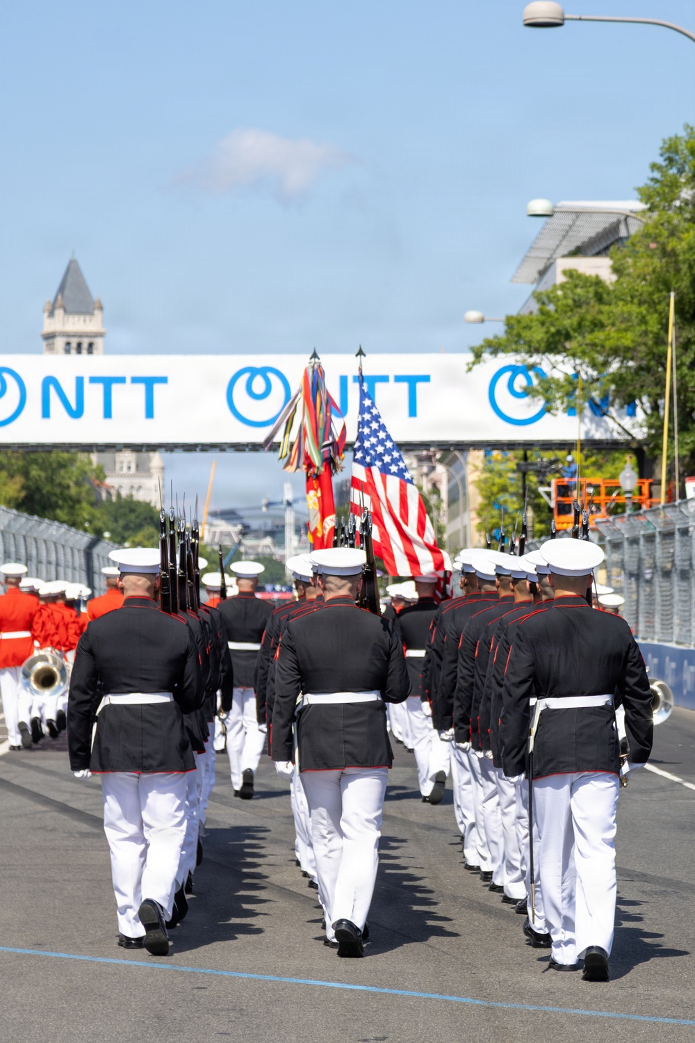 A U.S. Marine Corps ceremonial unit marches in formation down a street, carrying the American flag and military colors. The procession passes under an NTT-branded overhead banner, with a Marine Corps band in red uniforms visible to the left. The setting appears to be Washington, D.C., based on the visible architecture including what appears to be the Old Post Office Tower in the background.