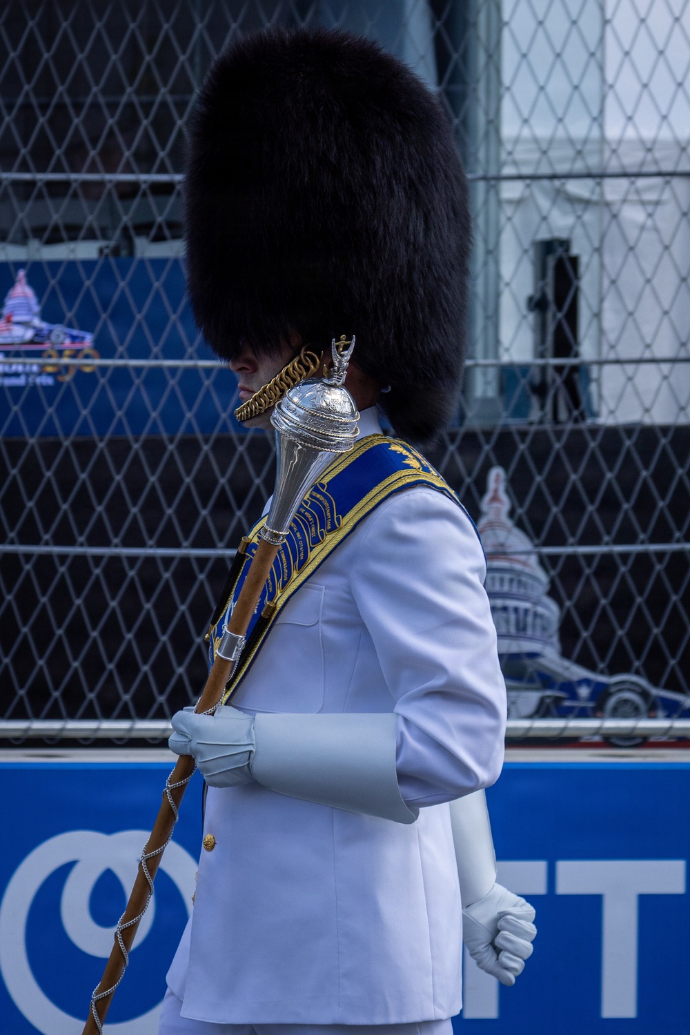 A marching band drum major or color guard member is shown in profile, wearing a tall black bearskin hat and a white uniform with a blue and gold sash. They are holding a ceremonial mace with a silver finial. The setting appears to be an outdoor event, possibly an IndyCar or Formula E race based on visible signage and a race car visible in the background.