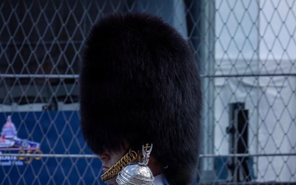 A marching band drum major or color guard member is shown in profile, wearing a tall black bearskin hat and a white uniform with a blue and gold sash. They are holding a ceremonial mace with a silver finial. The setting appears to be an outdoor event, possibly an IndyCar or Formula E race based on visible signage and a race car visible in the background.