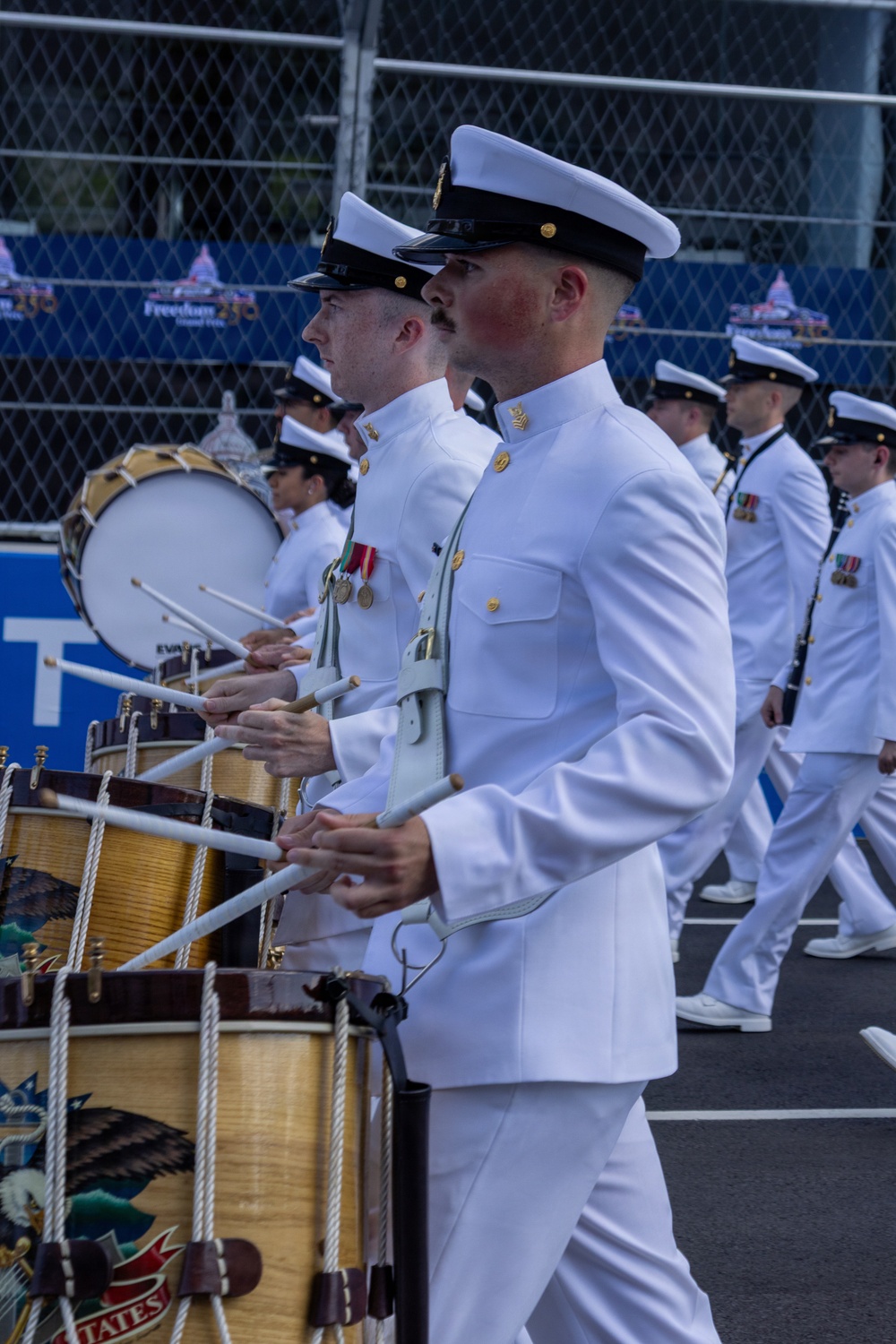 A United States Navy marching band is performing in a parade or ceremonial event. The drummers are dressed in formal white dress uniforms and are playing snare drums and a bass drum while marching in formation. A banner in the background references 'Freedom 250,' suggesting this is a patriotic or commemorative event.