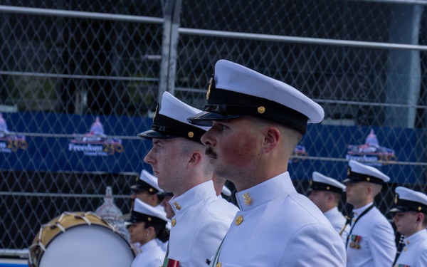 A United States Navy marching band is performing in a parade or ceremonial event. The drummers are dressed in formal white dress uniforms and are playing snare drums and a bass drum while marching in formation. A banner in the background references 'Freedom 250,' suggesting this is a patriotic or commemorative event.