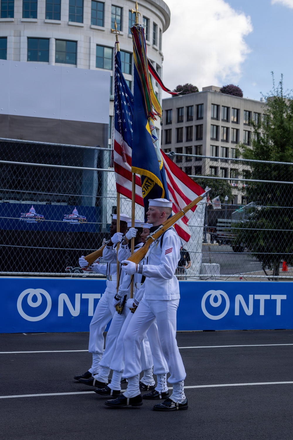 A U.S. Navy color guard is marching in formation carrying flags and rifles at what appears to be the Freedom Grand Prix event. The sailors are dressed in formal white dress uniforms and are performing a ceremonial presentation on a racing circuit. NTT sponsorship banners are prominently displayed along the track barrier in the background.