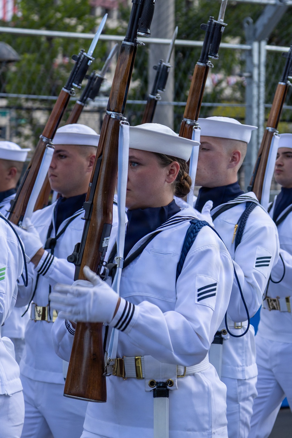 A formation of U.S. Navy personnel in dress white uniforms marching in a ceremonial parade or drill. The sailors are carrying rifles with fixed bayonets at shoulder arms position, wearing traditional white Dixie cup covers and white gloves. The formation appears disciplined and precise, consistent with a formal honor guard or fleet week-type public ceremony.