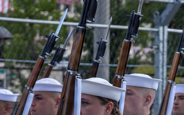 A formation of U.S. Navy personnel in dress white uniforms marching in a ceremonial parade or drill. The sailors are carrying rifles with fixed bayonets at shoulder arms position, wearing traditional white Dixie cup covers and white gloves. The formation appears disciplined and precise, consistent with a formal honor guard or fleet week-type public ceremony.