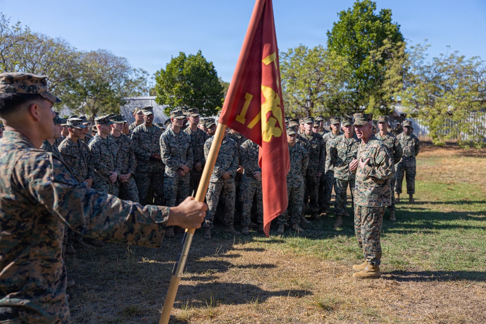 A senior military officer addresses a formation of U.S. Marines in an outdoor setting. A guidon bearer holds a red Marine Corps unit flag in the foreground while the assembled Marines stand at ease listening. The setting appears to be a grassy field with trees and a structure visible in the background.