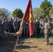 A senior military officer addresses a formation of U.S. Marines in an outdoor setting. A guidon bearer holds a red Marine Corps unit flag in the foreground while the assembled Marines stand at ease listening. The setting appears to be a grassy field with trees and a structure visible in the background.