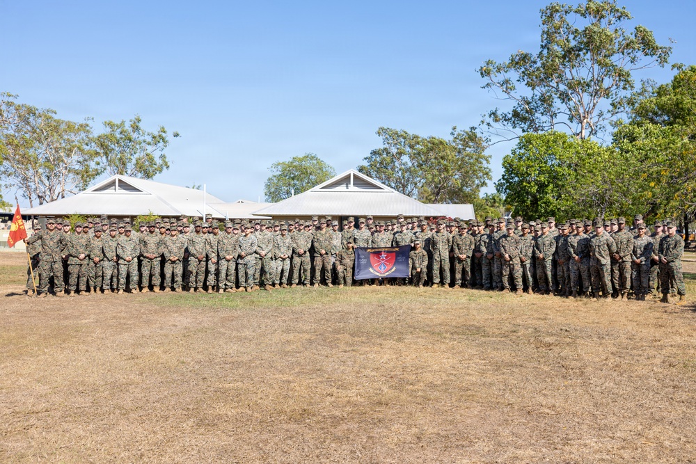 A large group of military personnel, appearing to be U.S. Marines based on their woodland MARPAT camouflage uniforms, are posed for a formal unit photograph outdoors. They are arranged in multiple rows on a dry grass field, with low-profile buildings and eucalyptus trees visible in the background, suggesting a location in Australia. A unit guidon flag and a dark banner with a red shield insignia are visible among the group.