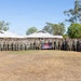 A large group of military personnel, appearing to be U.S. Marines based on their woodland MARPAT camouflage uniforms, are posed for a formal unit photograph outdoors. They are arranged in multiple rows on a dry grass field, with low-profile buildings and eucalyptus trees visible in the background, suggesting a location in Australia. A unit guidon flag and a dark banner with a red shield insignia are visible among the group.