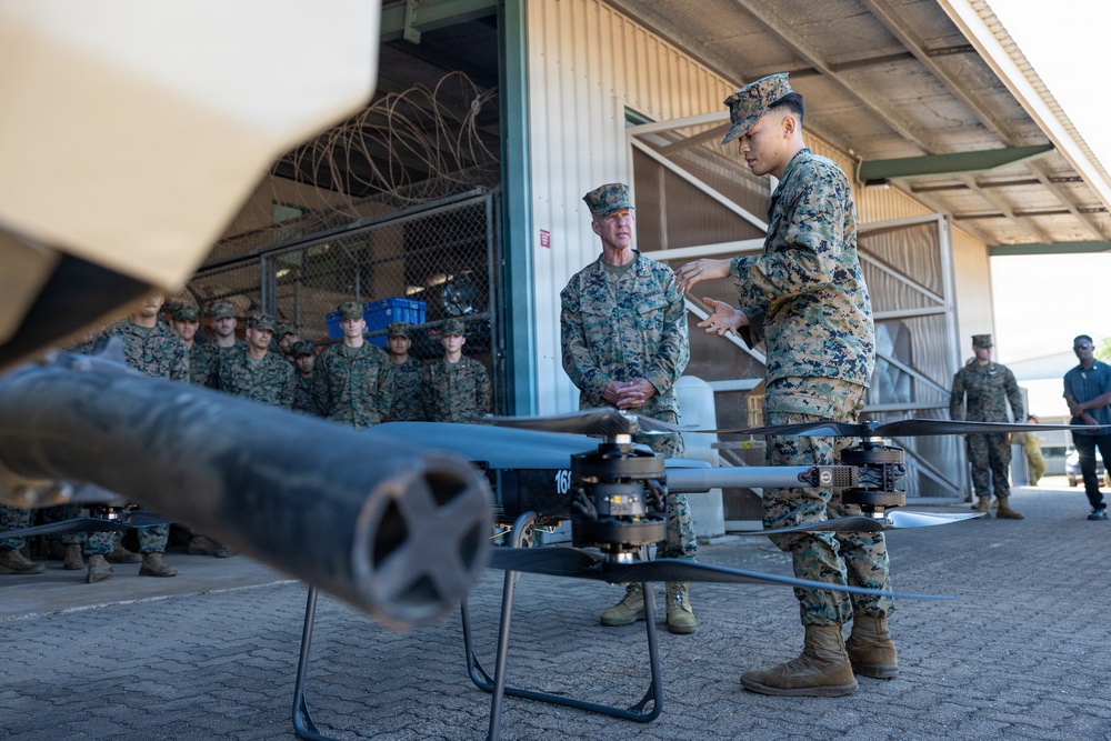 Military personnel in MARPAT camouflage uniforms are gathered at what appears to be a military installation for a briefing or demonstration involving an armed drone/UAS (Unmanned Aerial System). A junior Marine appears to be presenting or explaining the drone to a senior officer while a group of Marines observes in the background. The drone in the foreground is equipped with what appears to be a weapon system or launcher mounted on its frame.