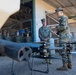 Military personnel in MARPAT camouflage uniforms are gathered at what appears to be a military installation for a briefing or demonstration involving an armed drone/UAS (Unmanned Aerial System). A junior Marine appears to be presenting or explaining the drone to a senior officer while a group of Marines observes in the background. The drone in the foreground is equipped with what appears to be a weapon system or launcher mounted on its frame.