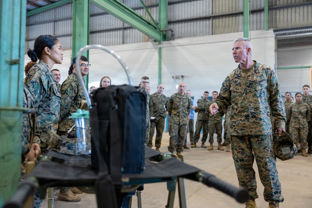A senior military officer in U.S. Marine Corps MARPAT camouflage uniform addresses a large group of Marines inside an industrial hangar facility. The officer, holding a cover (hat) in his left hand, appears to be conducting a briefing or demonstration near a piece of equipment on a stand. A female Marine stands opposite him, likely presenting or explaining the equipment.
