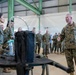 A senior military officer in U.S. Marine Corps MARPAT camouflage uniform addresses a large group of Marines inside an industrial hangar facility. The officer, holding a cover (hat) in his left hand, appears to be conducting a briefing or demonstration near a piece of equipment on a stand. A female Marine stands opposite him, likely presenting or explaining the equipment.
