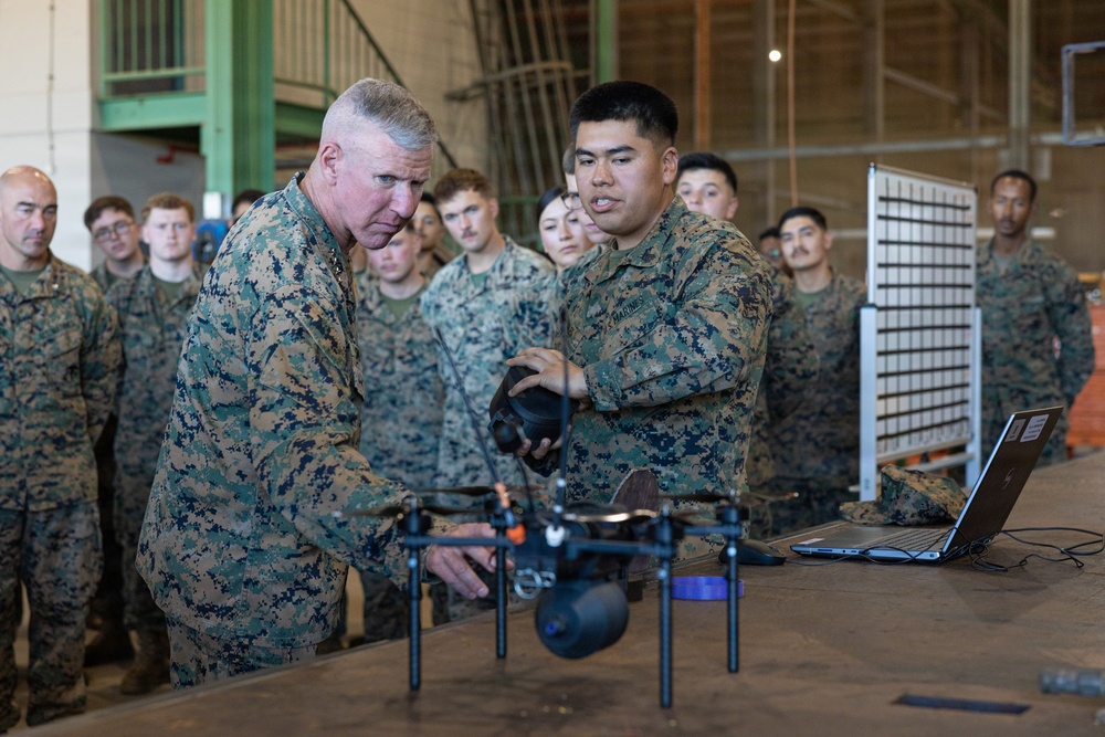 Two U.S. Marines in MARPAT camouflage uniforms are examining a small multi-rotor drone on a table inside what appears to be a military hangar or warehouse facility. A senior officer (identifiable by stars on collar) is being briefed by a junior Marine on the drone's capabilities, with a laptop computer nearby. A group of approximately 15-20 military personnel observe the demonstration in the background.