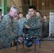 Two U.S. Marines in MARPAT camouflage uniforms are examining a small multi-rotor drone on a table inside what appears to be a military hangar or warehouse facility. A senior officer (identifiable by stars on collar) is being briefed by a junior Marine on the drone's capabilities, with a laptop computer nearby. A group of approximately 15-20 military personnel observe the demonstration in the background.