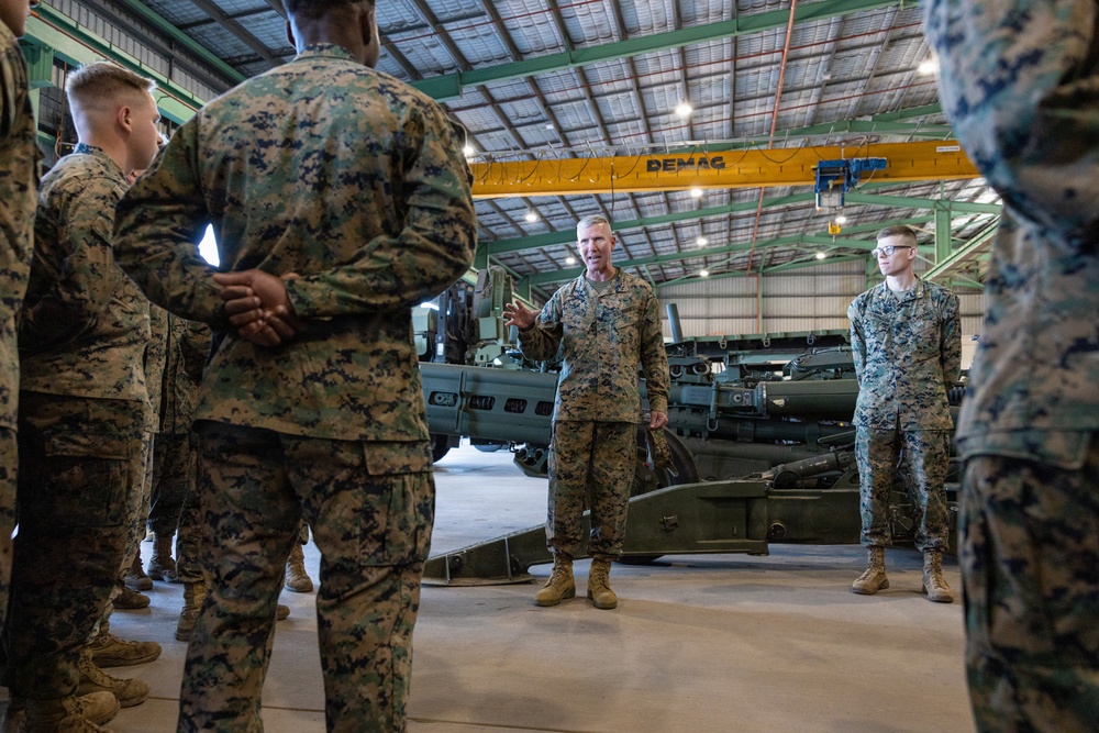 A group of military personnel in MARPAT (Marine pattern) camouflage uniforms are gathered inside a large industrial hangar. A senior service member appears to be addressing or briefing the group, standing in the center while others listen. Military equipment, including what appears to be artillery pieces, is visible in the background.