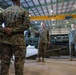 A group of military personnel in MARPAT (Marine pattern) camouflage uniforms are gathered inside a large industrial hangar. A senior service member appears to be addressing or briefing the group, standing in the center while others listen. Military equipment, including what appears to be artillery pieces, is visible in the background.