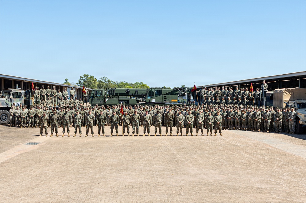 A large formation of military personnel, appearing to be U.S. Marines based on their camouflage uniforms and unit guidons, pose for a group photograph in a motor pool or parade ground area. The formation is arranged in multiple rows with military vehicles and equipment visible in the background, flanked by open-bay maintenance or storage structures. Several red and gold unit guidons (flags) are visible, consistent with U.S. Marine Corps colors.