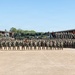 A large formation of military personnel, appearing to be U.S. Marines based on their camouflage uniforms and unit guidons, pose for a group photograph in a motor pool or parade ground area. The formation is arranged in multiple rows with military vehicles and equipment visible in the background, flanked by open-bay maintenance or storage structures. Several red and gold unit guidons (flags) are visible, consistent with U.S. Marine Corps colors.