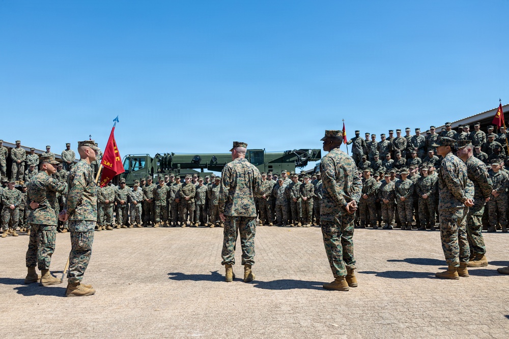 A formal U.S. Marine Corps military ceremony is taking place on an outdoor parade deck or drill field. Several Marines in leadership positions stand facing a large formation of troops arranged in ranks. Red guidon flags and military vehicles are visible in the background.