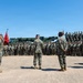 A formal U.S. Marine Corps military ceremony is taking place on an outdoor parade deck or drill field. Several Marines in leadership positions stand facing a large formation of troops arranged in ranks. Red guidon flags and military vehicles are visible in the background.