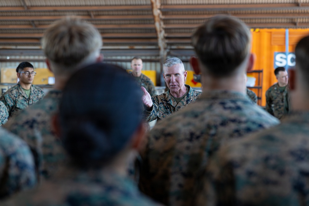 A senior military officer in U.S. Marine Corps MARPAT camouflage uniform addresses a group of Marines in what appears to be a hangar or industrial facility. The central figure, who has gray hair and visible rank insignia, is gesturing with his hand while speaking to the assembled personnel. The Marines are gathered in a semi-circle facing the speaker, suggesting a briefing or command address.