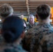 A senior military officer in U.S. Marine Corps MARPAT camouflage uniform addresses a group of Marines in what appears to be a hangar or industrial facility. The central figure, who has gray hair and visible rank insignia, is gesturing with his hand while speaking to the assembled personnel. The Marines are gathered in a semi-circle facing the speaker, suggesting a briefing or command address.