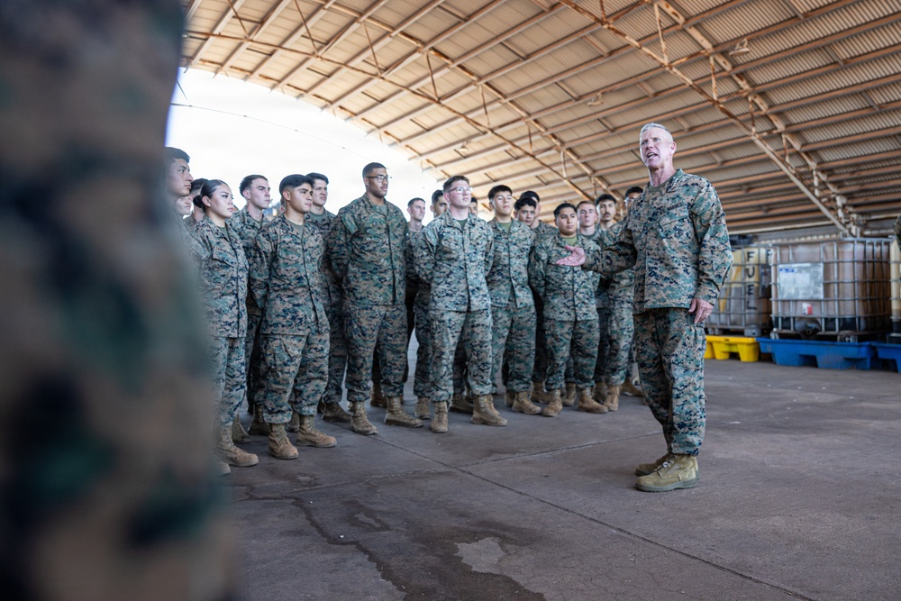 A senior military officer in MARPAT camouflage uniform addresses a formation of younger service members inside a large covered warehouse or hangar facility. The officer appears to be speaking or briefing the assembled group, who are standing at ease in a semi-formal formation. The setting suggests a military base or logistics facility.