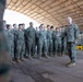 A senior military officer in MARPAT camouflage uniform addresses a formation of younger service members inside a large covered warehouse or hangar facility. The officer appears to be speaking or briefing the assembled group, who are standing at ease in a semi-formal formation. The setting suggests a military base or logistics facility.