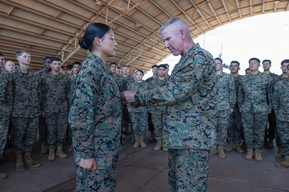 A senior military officer in US Marine Corps MARPAT digital camouflage uniform is pinning a medal or award onto a junior female service member during a formal ceremony. The event takes place inside a large open-sided hangar or shelter, with a formation of Marines standing at attention in the background. This appears to be an official military awards or recognition ceremony.