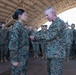 A senior military officer in US Marine Corps MARPAT digital camouflage uniform is pinning a medal or award onto a junior female service member during a formal ceremony. The event takes place inside a large open-sided hangar or shelter, with a formation of Marines standing at attention in the background. This appears to be an official military awards or recognition ceremony.