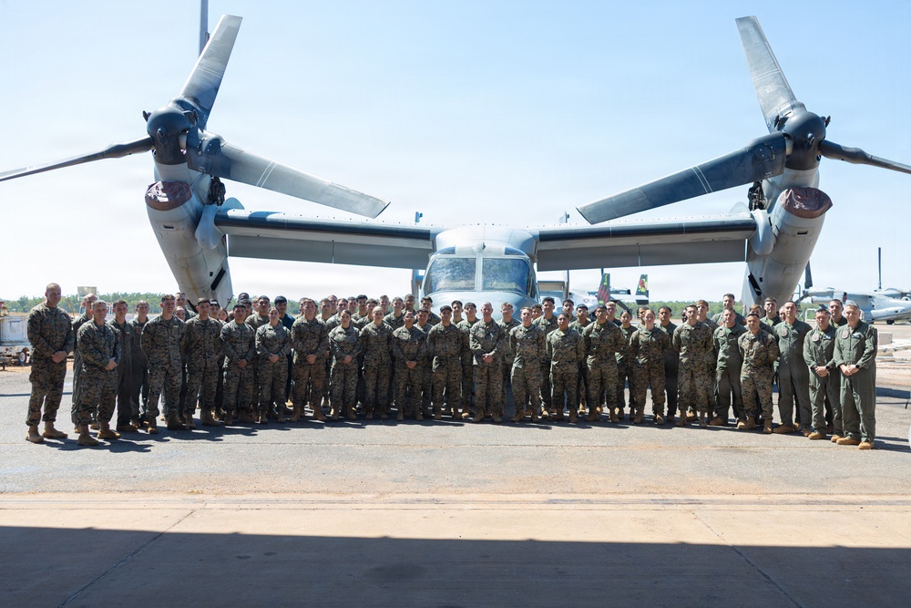 A large group of military personnel, predominantly in Marine Corps MARPAT camouflage uniforms with a smaller number in flight suits, pose for a formal group photograph on a flight line tarmac. They are standing in front of a V-22 Osprey tiltrotor aircraft with its rotors in the horizontal (airplane mode) position. The setting appears to be a military air station on a clear, sunny day.